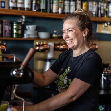 Kvinde står ved en kaffemaskine i en cafe og smiler med flasker i baggrunden 